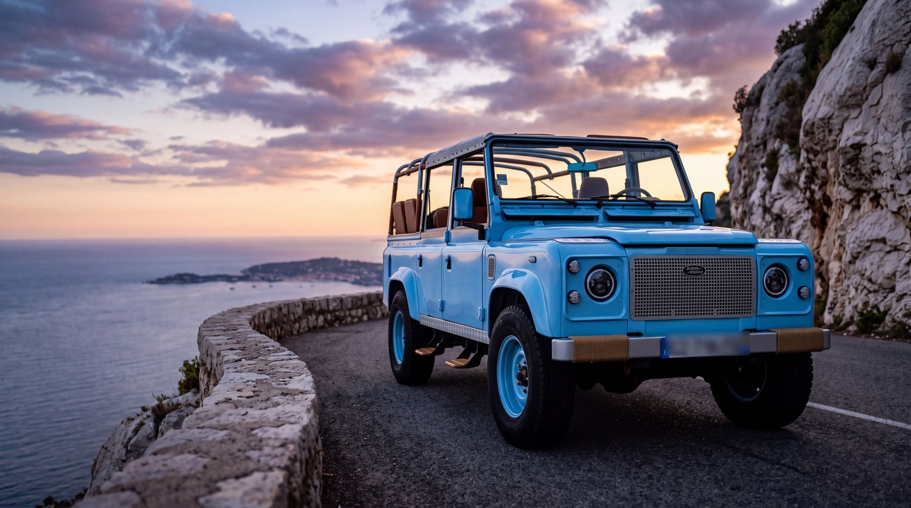 Land Rover Defender bleu ciel sur une route en corniche au crépuscule, surplombant la Méditerranée